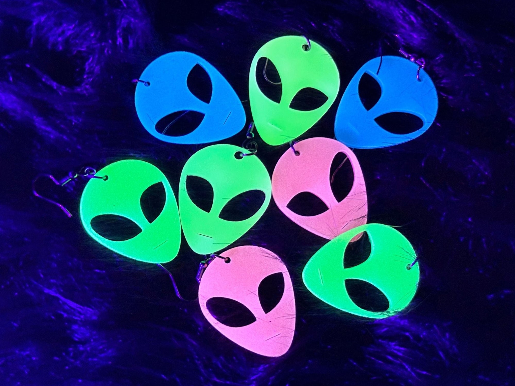 Glow in the Dark alien head earrings in multiple acrylic colors arranged on faux fur background in blacklight.