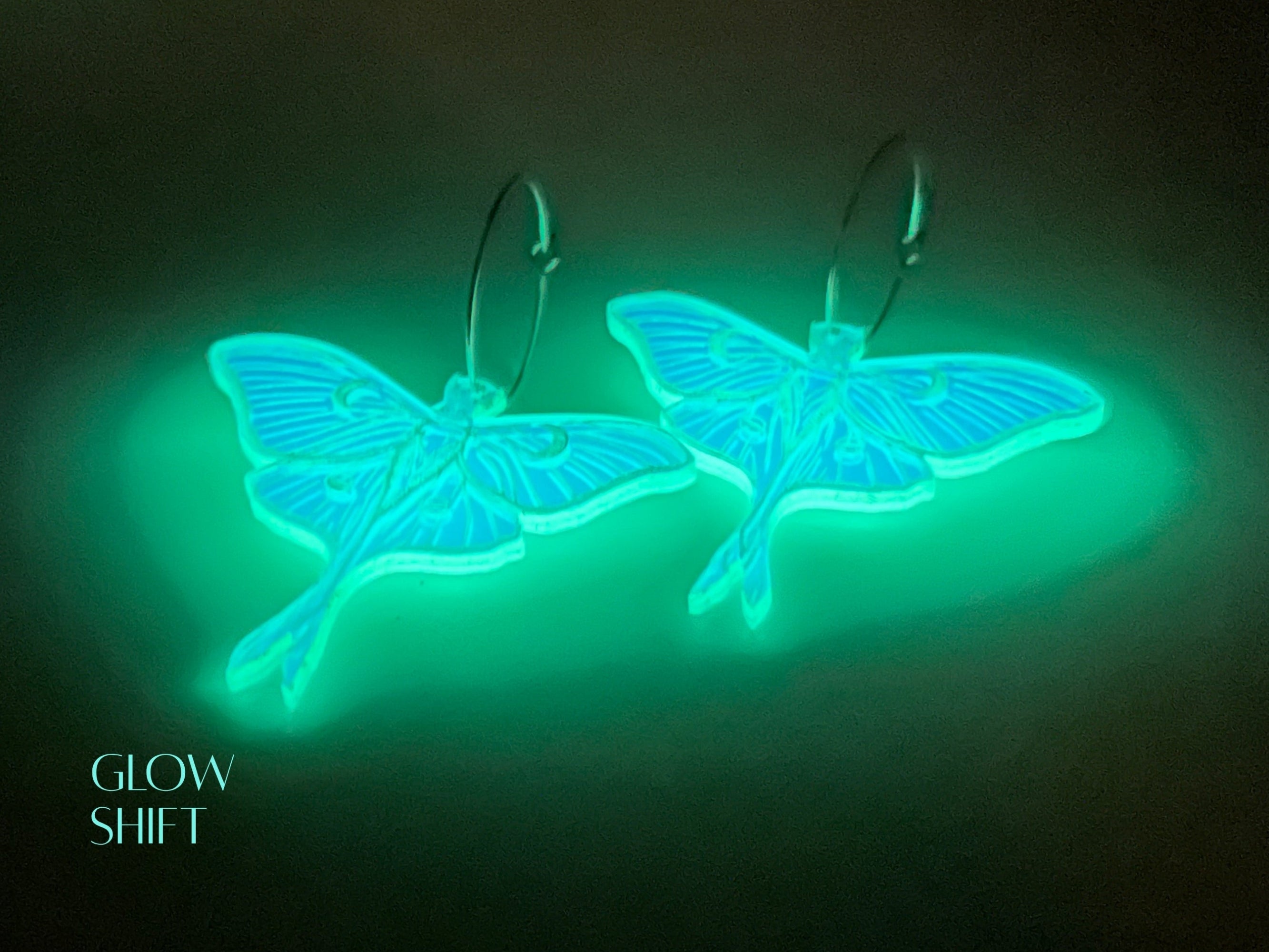 Pair of Luna moth earrings glowing bright aqua under low light, demonstrating the luminous appearance of the acrylic.