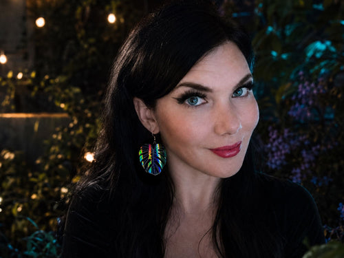 Woman wearing black rainbow iridescent monstera leaf statement earrings.