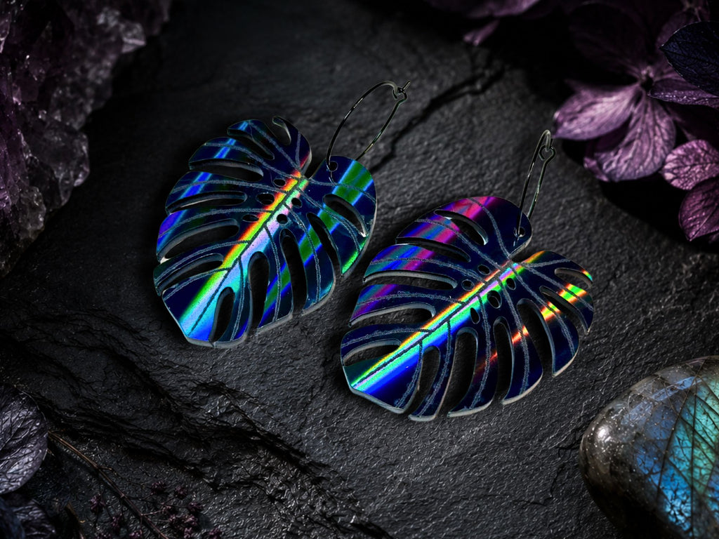 air of black rainbow iridescent monstera leaf earrings on a dark stone background.