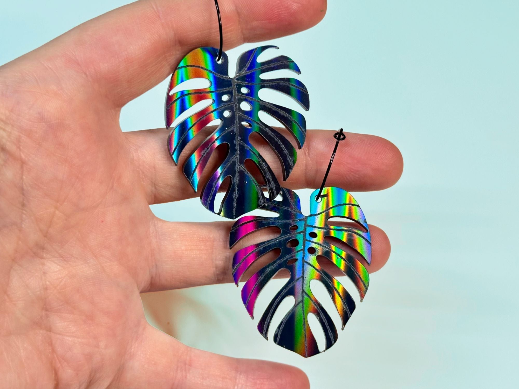Hand holding black rainbow iridescent monstera leaf earrings to show size.