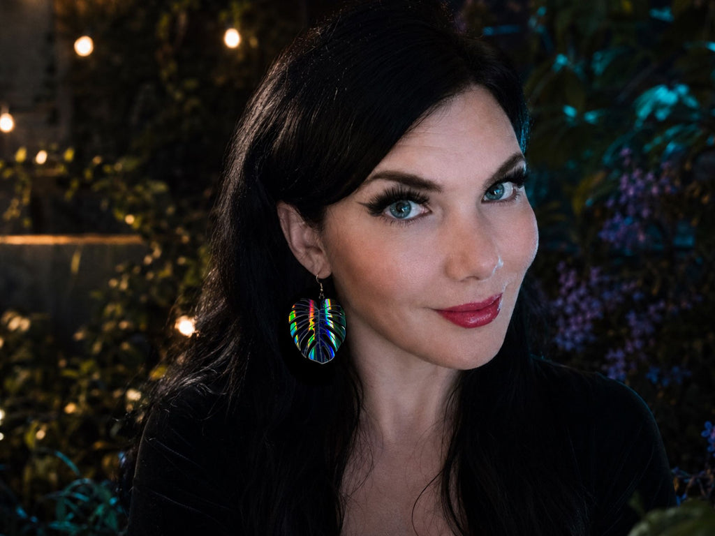 Woman wearing black rainbow iridescent monstera leaf statement earrings.