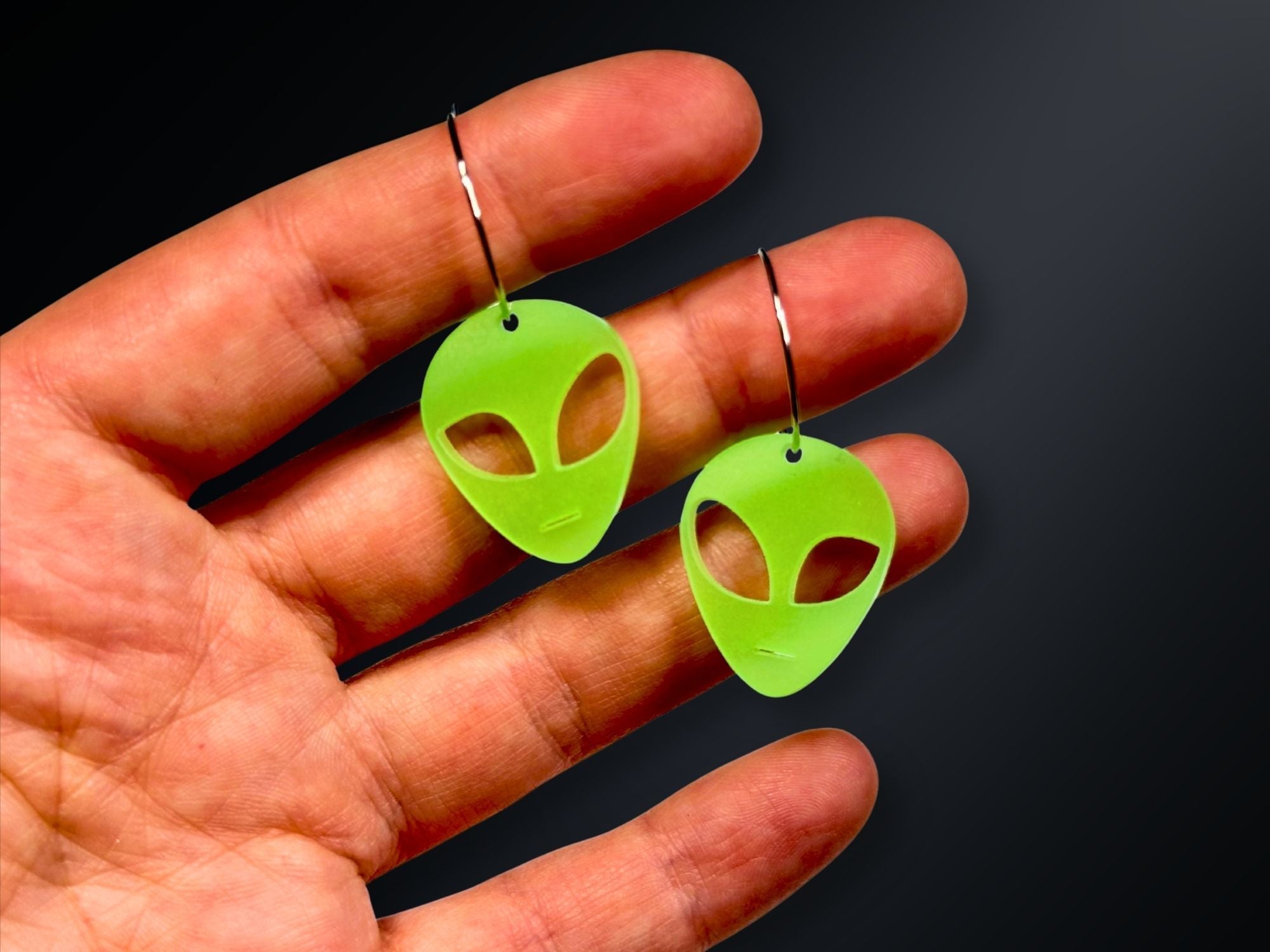 Hand holding smaller everyday size glow in the dark alien earrings in neon green acrylic.