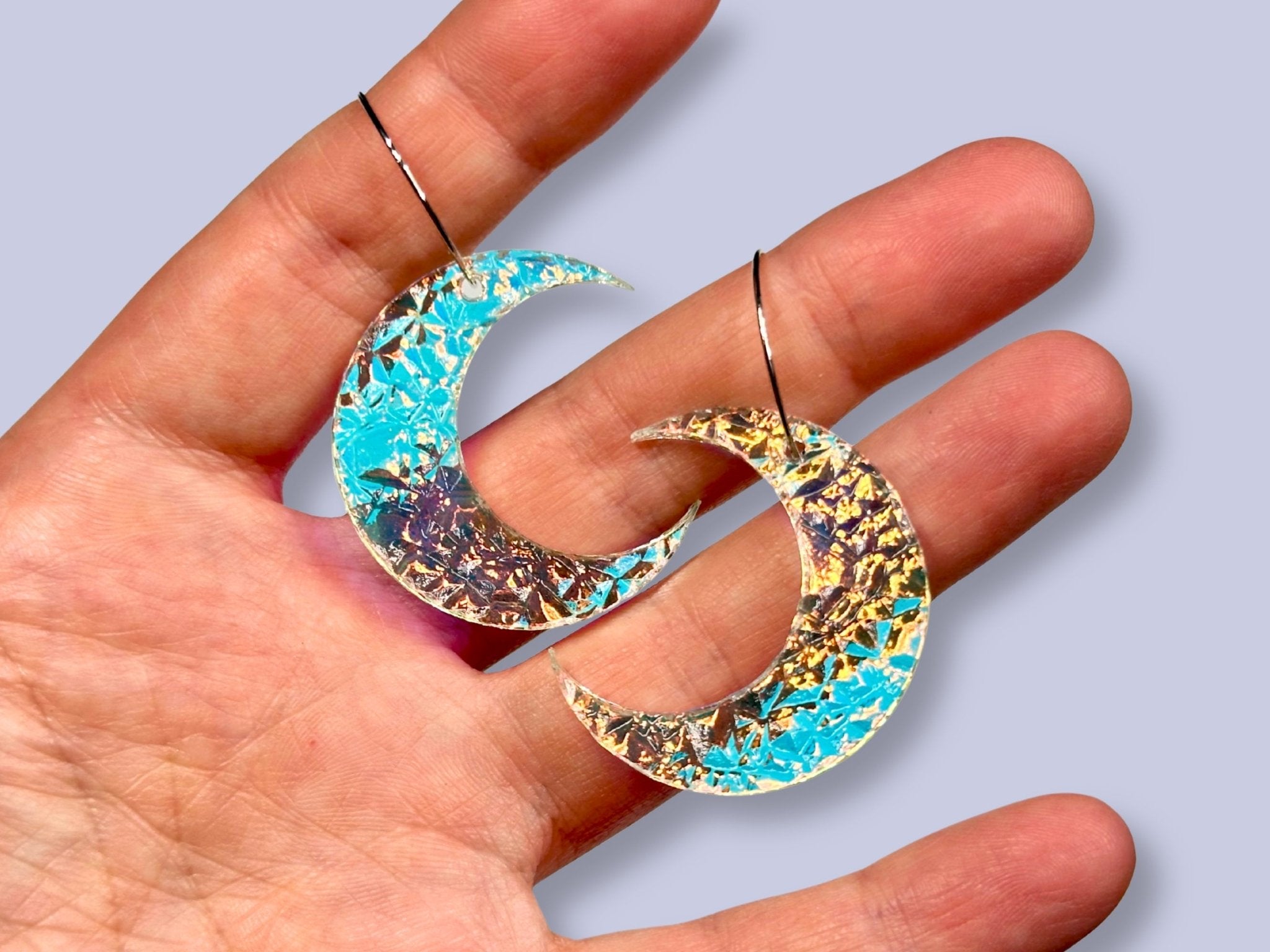 Hand holding two textured crescent moon earrings in turquoise-blue and gold iridescent finishes.