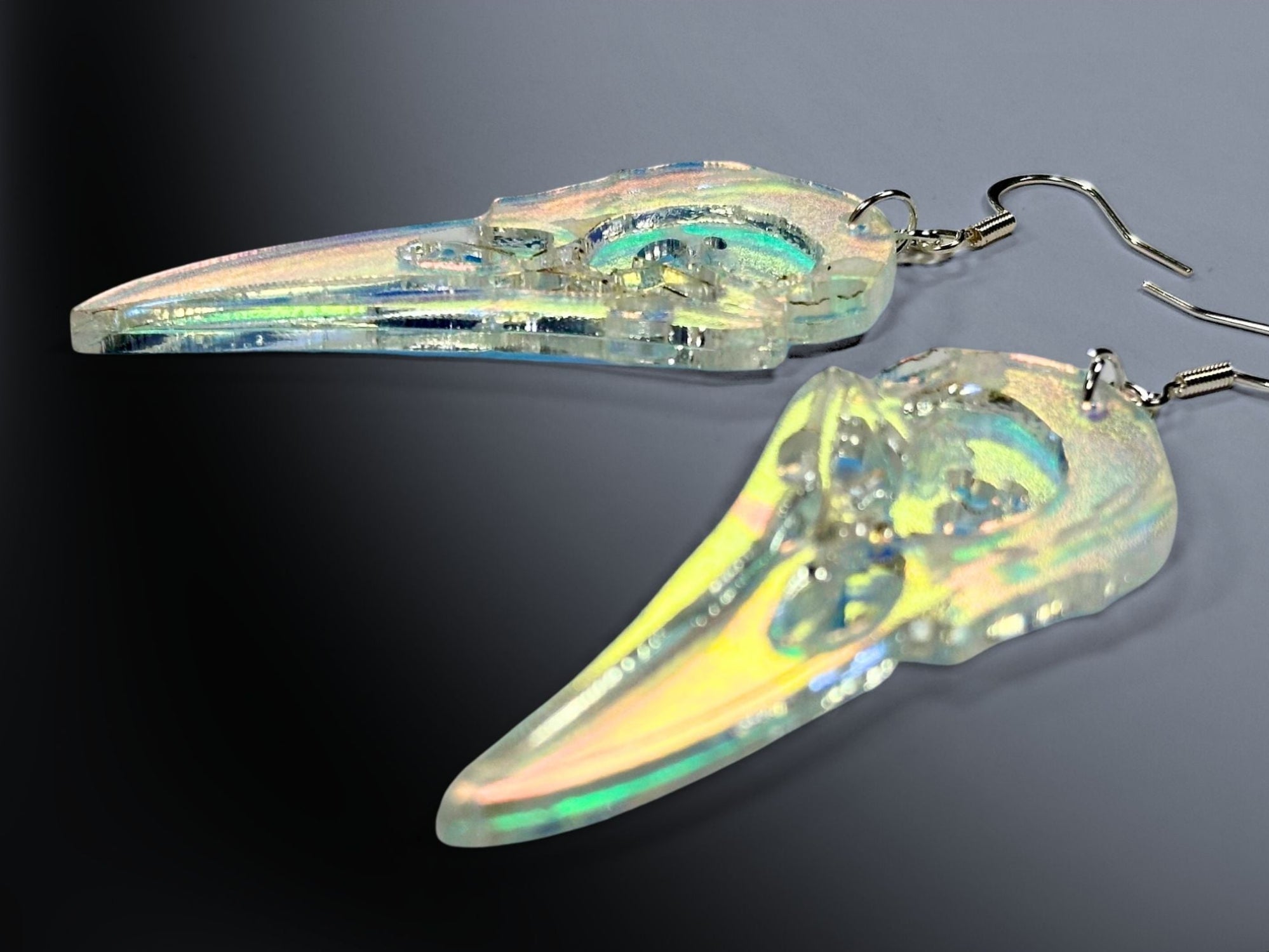 Close-up of raven skull statement earrings in translucent iridescent acrylic on dark gradient background.