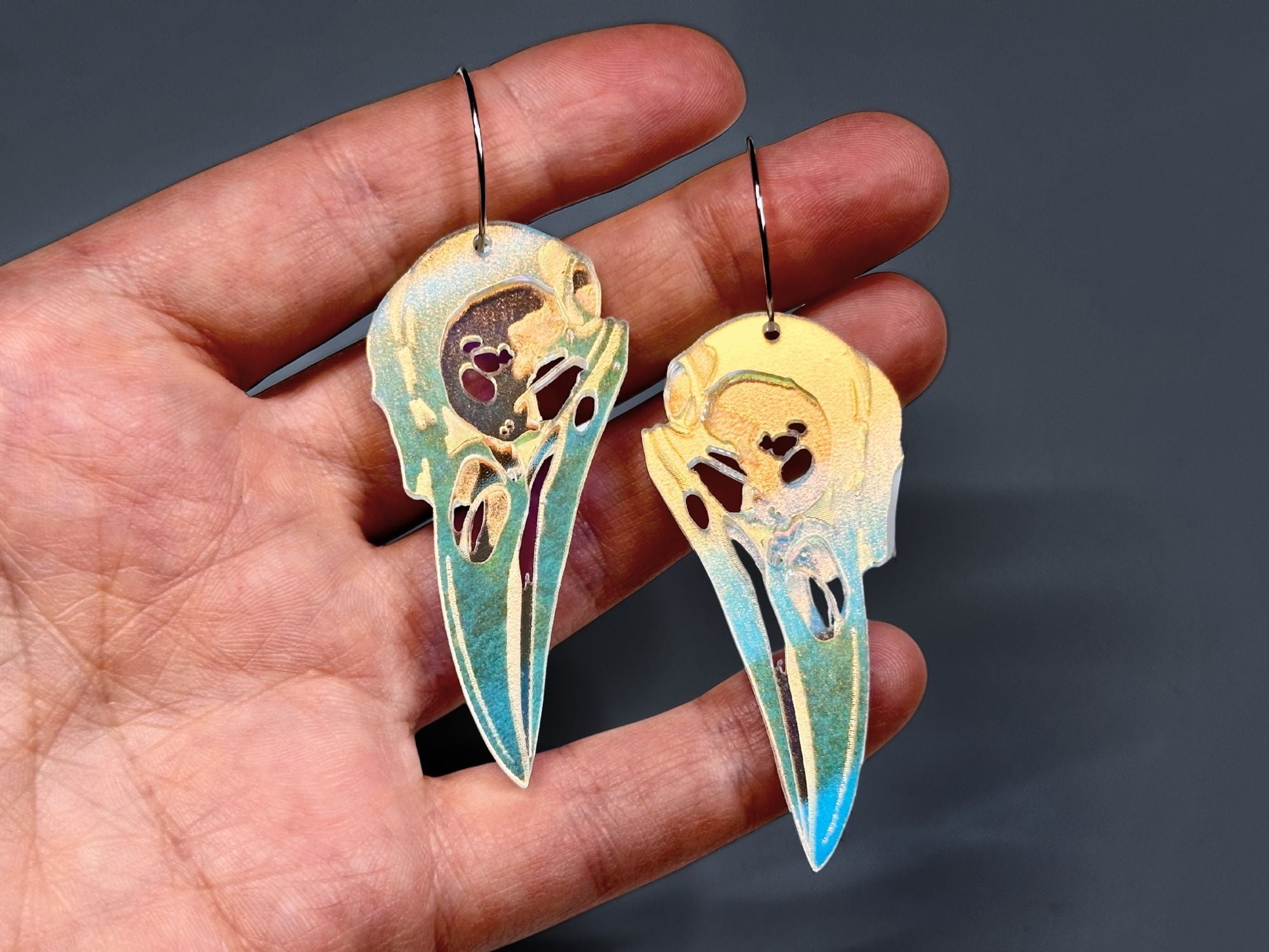 Hand holding iridescent raven skull acrylic earrings for scale reference.