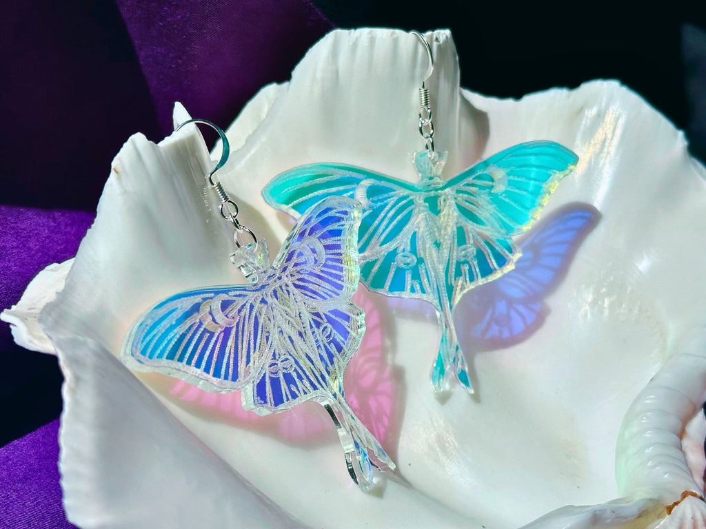 Two luna moth earrings with iridescent blue, purple, and turquoise wings, displayed on a white clam shell against a dark purple background.