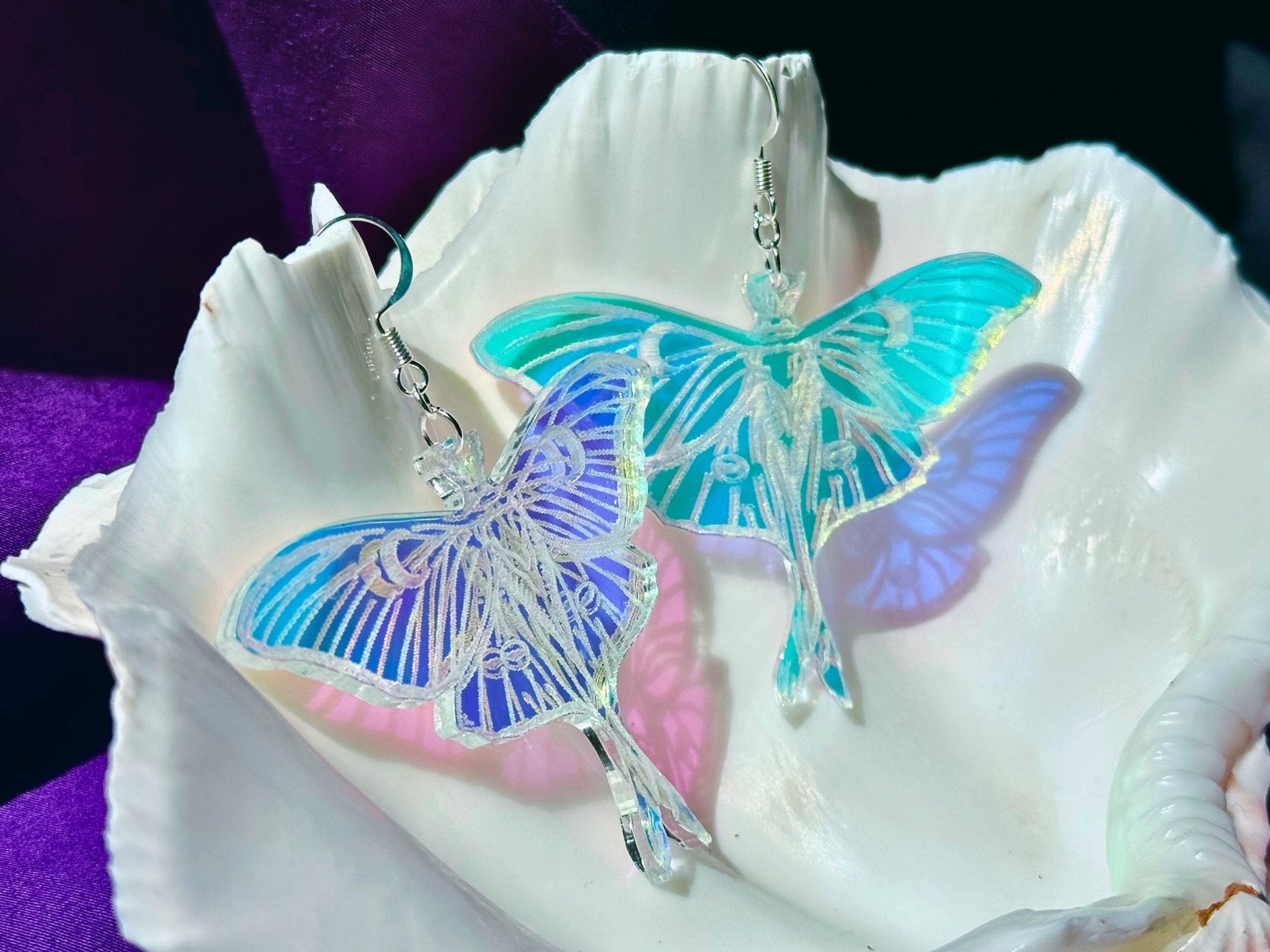 Two luna moth earrings with iridescent blue, purple, and turquoise wings, displayed on a white clam shell against a dark purple background.