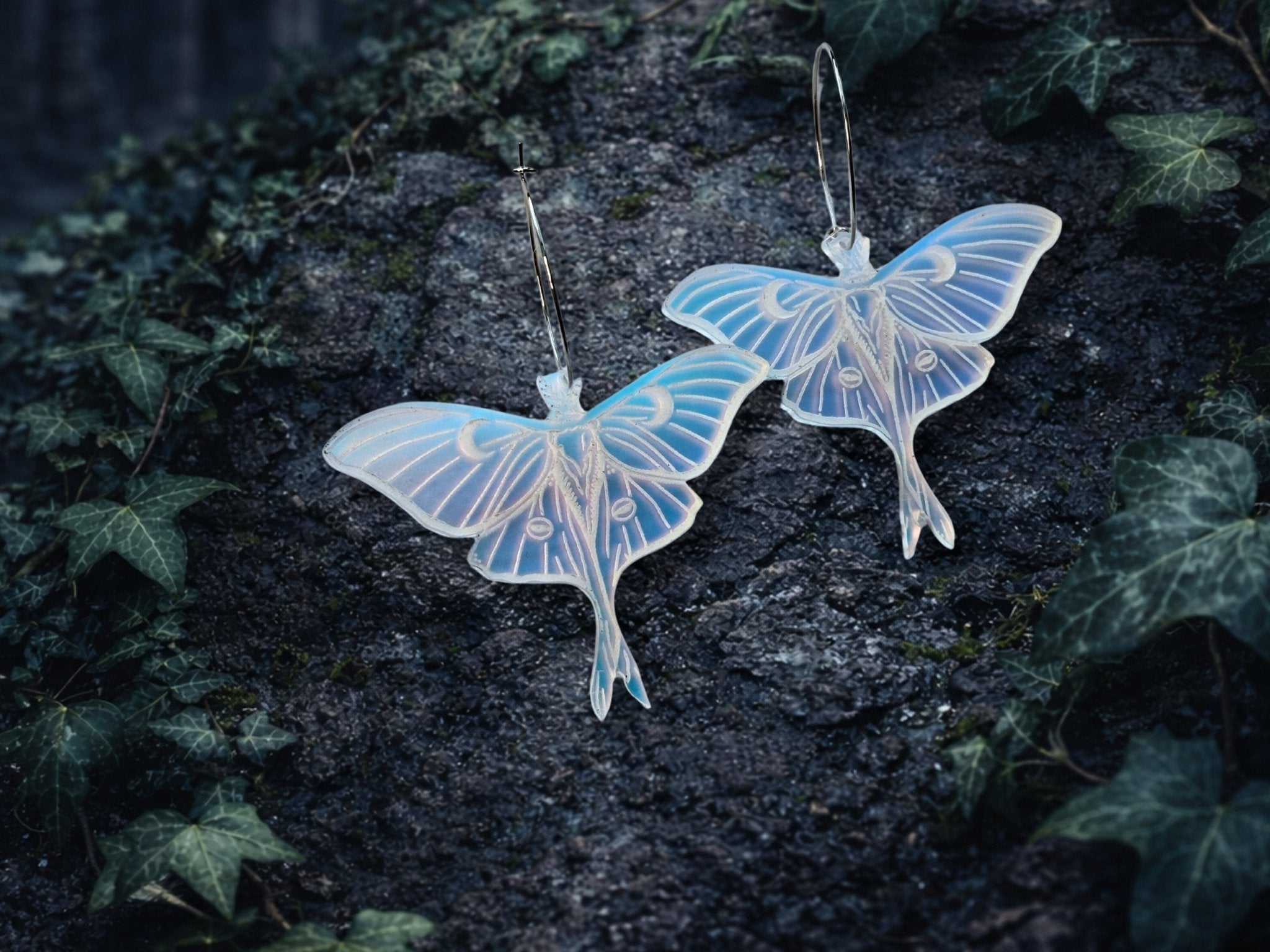 Pair of frosted iridescent Luna moth earrings photographed outdoors on dark stone, with pale blue and lavender color shifts.