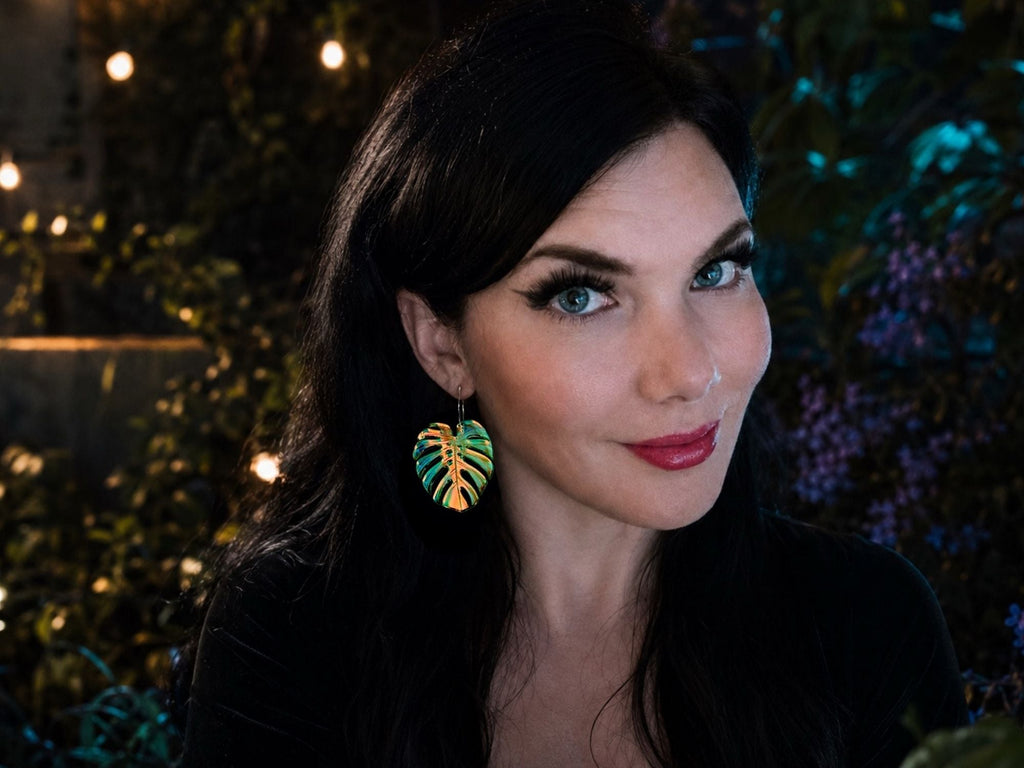 Woman wearing large iridescent monstera leaf statement earrings that shimmer in shades of blue, green, pink, and gold.
