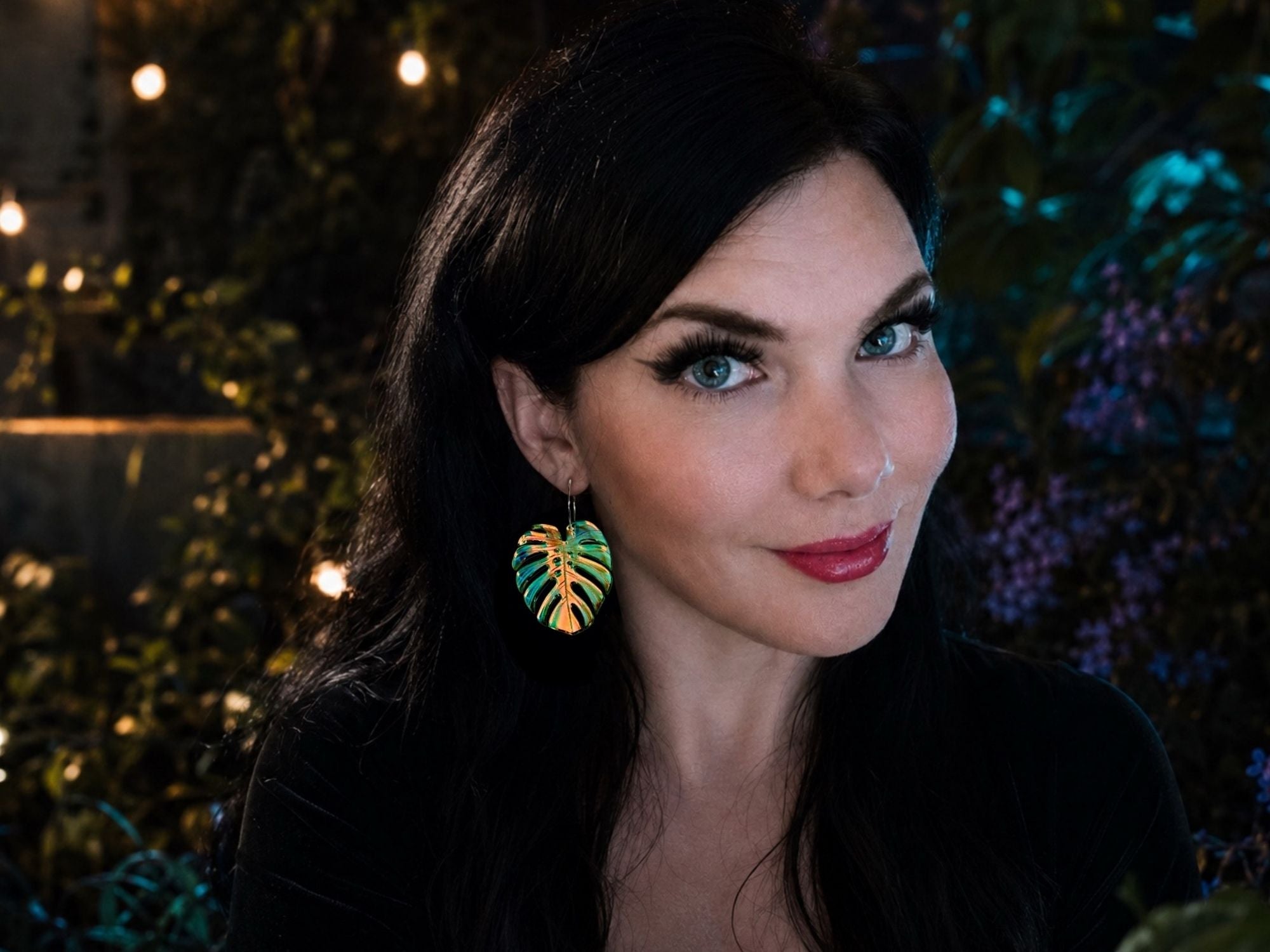 Woman wearing large iridescent monstera leaf statement earrings that shimmer in shades of blue, green, pink, and gold.