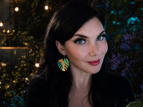 Woman wearing large iridescent monstera leaf statement earrings that shimmer in shades of blue, green, pink, and gold.