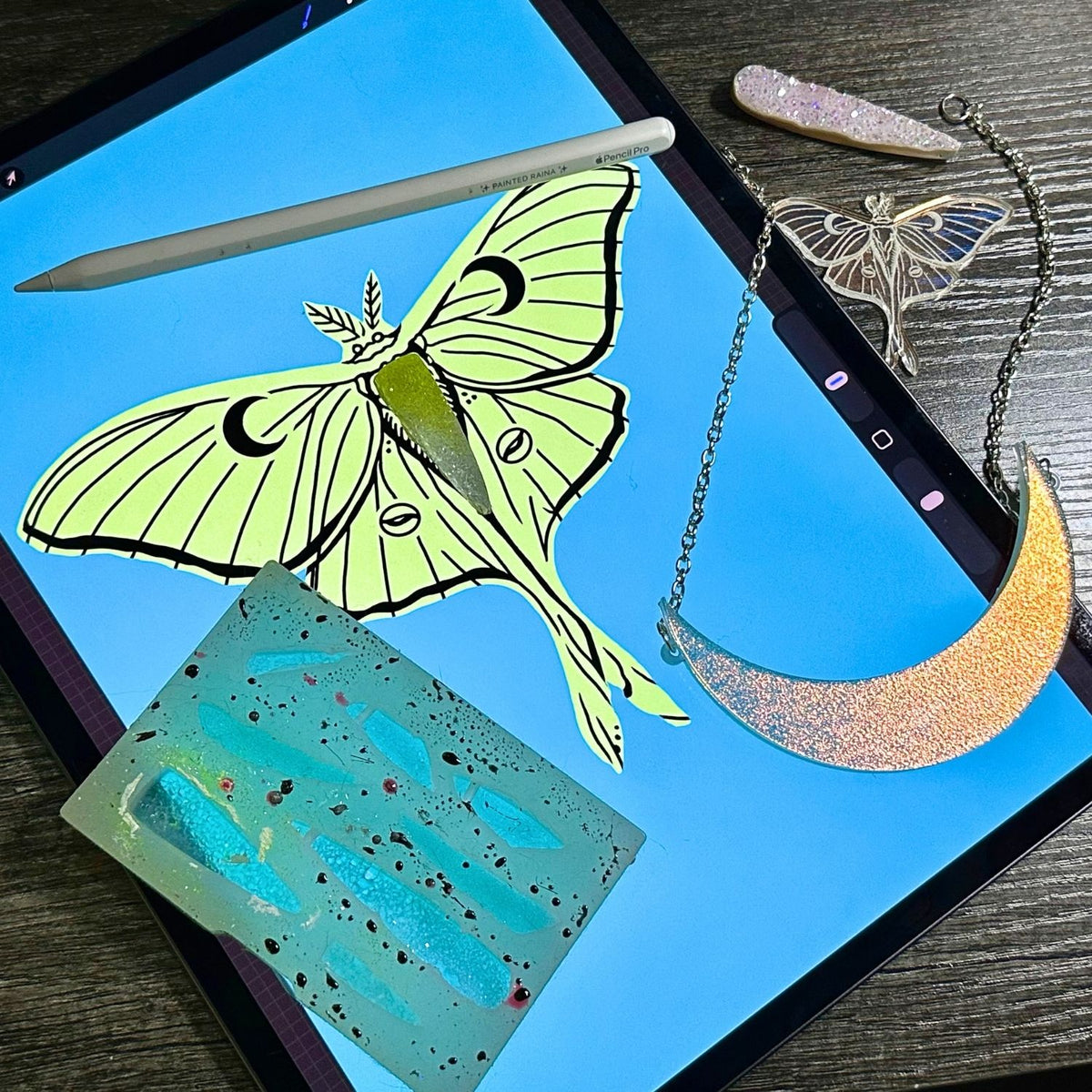 A styled flat-lay of jewelry and accessories arranged on an iPad screen over a dark wood surface. There is a crescent moon pendant necklace with a glittery gold-to-silver gradient shown alongside a resin mold, an Apple Pencil, a crystal, and a luna moth pendant.