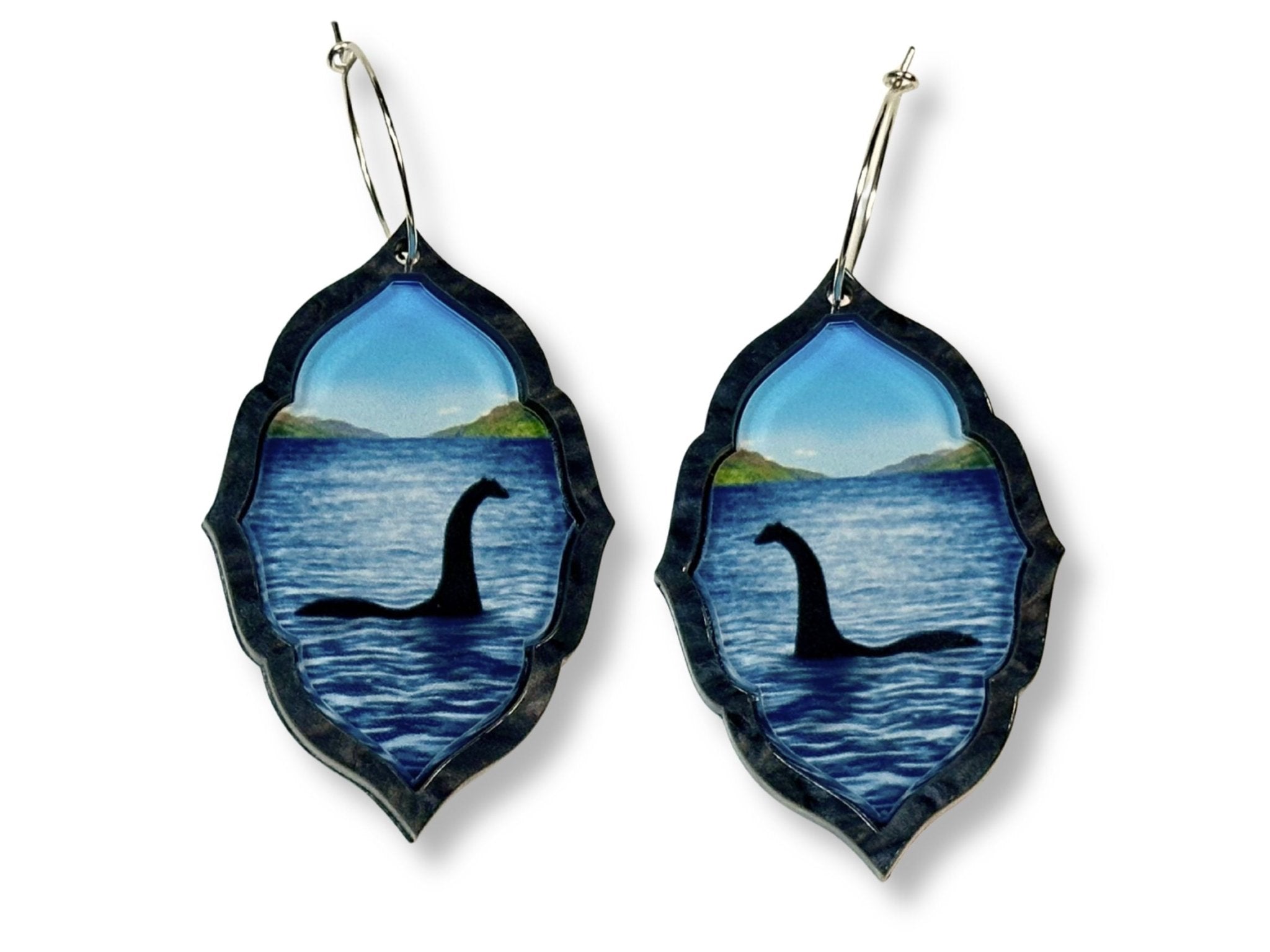Loch Ness monster statement earrings in layered acrylic with blue lake scene and silver hoop ear wires on white background.