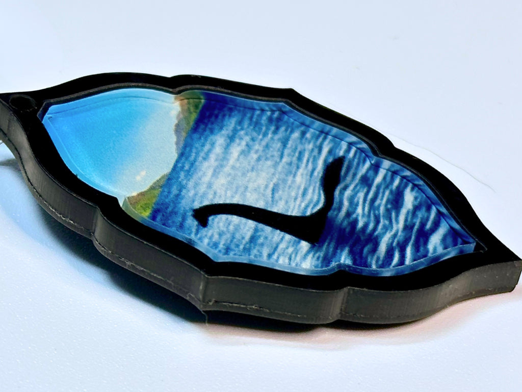 Close-up of original Loch Ness Monster pendant with blue lake artwork framed in black pearl acrylic.