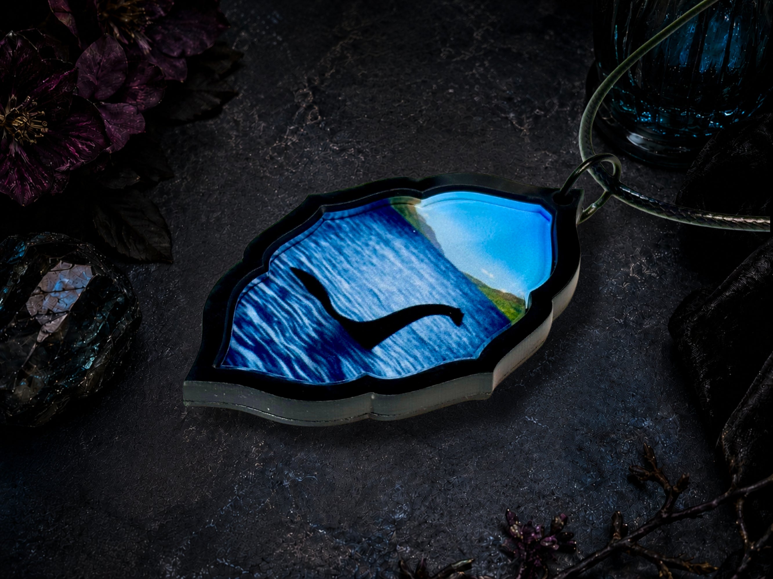 Angled flat lay of Loch Ness Monster pendant showing layered acrylic construction and blue water artwork.