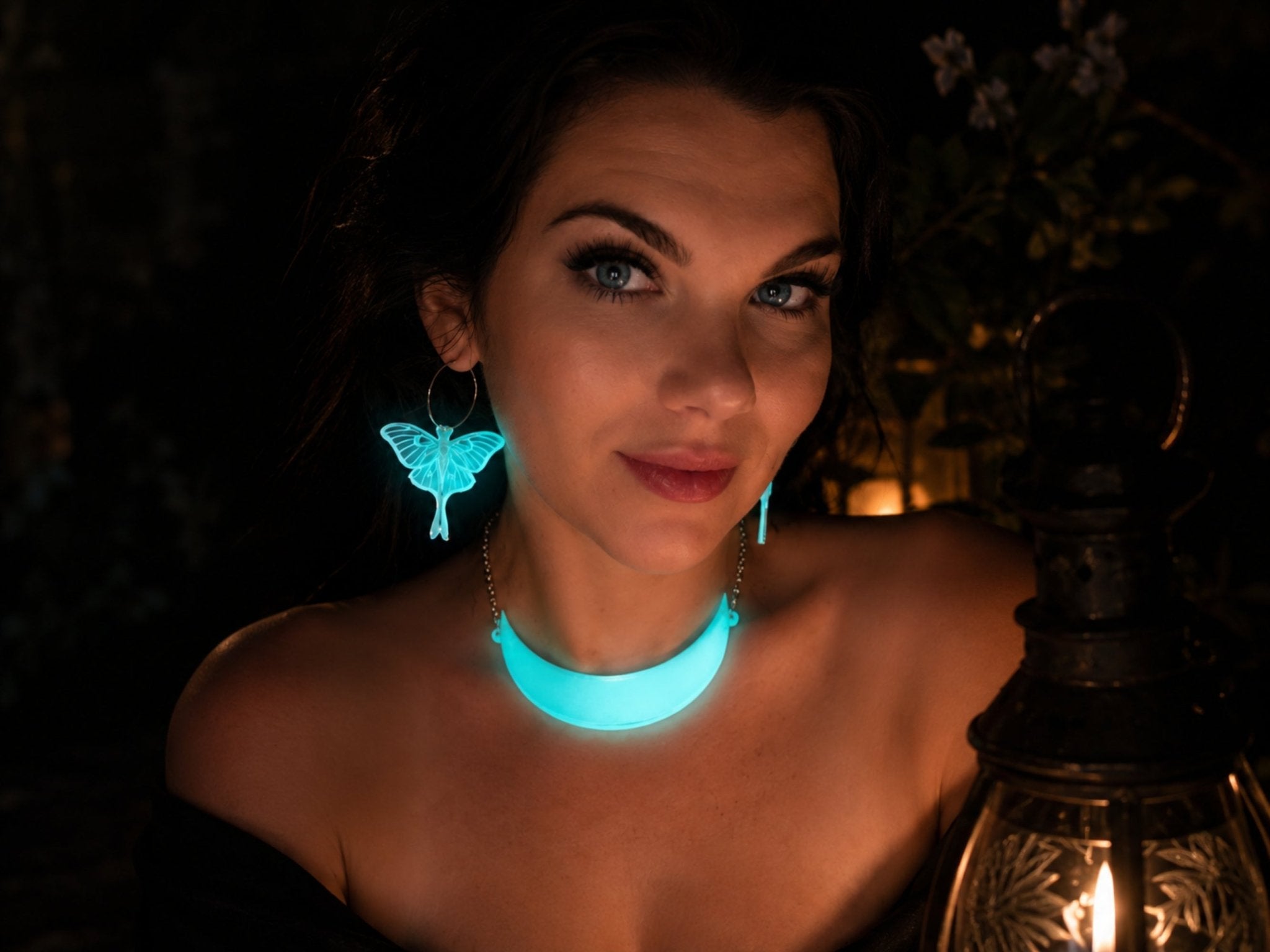 A woman wearing a Glow-in-the-dark crescent moon necklace and glowing luna moth hoop earrings, photographed in a dark setting with a lantern nearby. 