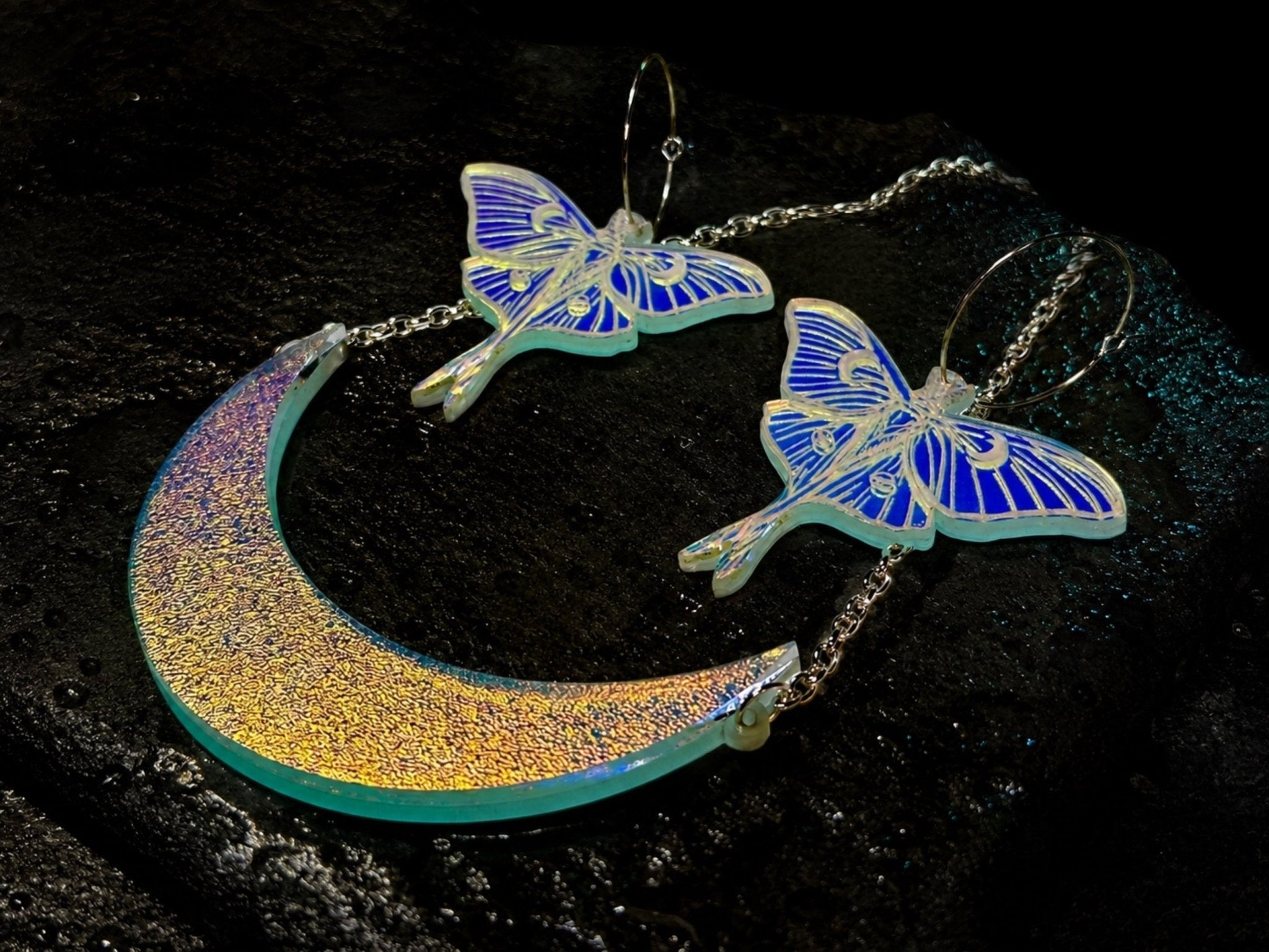 A pair of dangling earrings and a crescent moon pendant set on a dark textured surface. The crescent moon pendant has an iridescent gold-to-teal gradient finish, while the matching luna moth earrings show bright blue and white patterns and hang from silver-toned hoops.