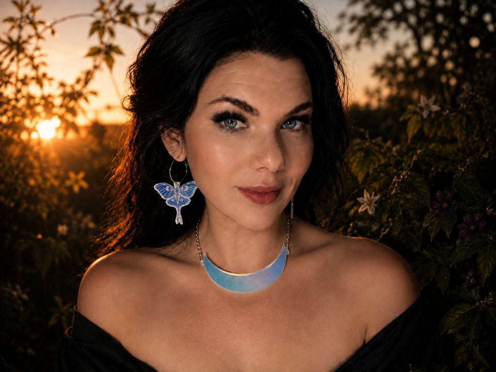 A woman outdoors at sunset wearing an Iridescent crescent moon collar necklace and luna moth earrings together, with dark foliage in the background.
