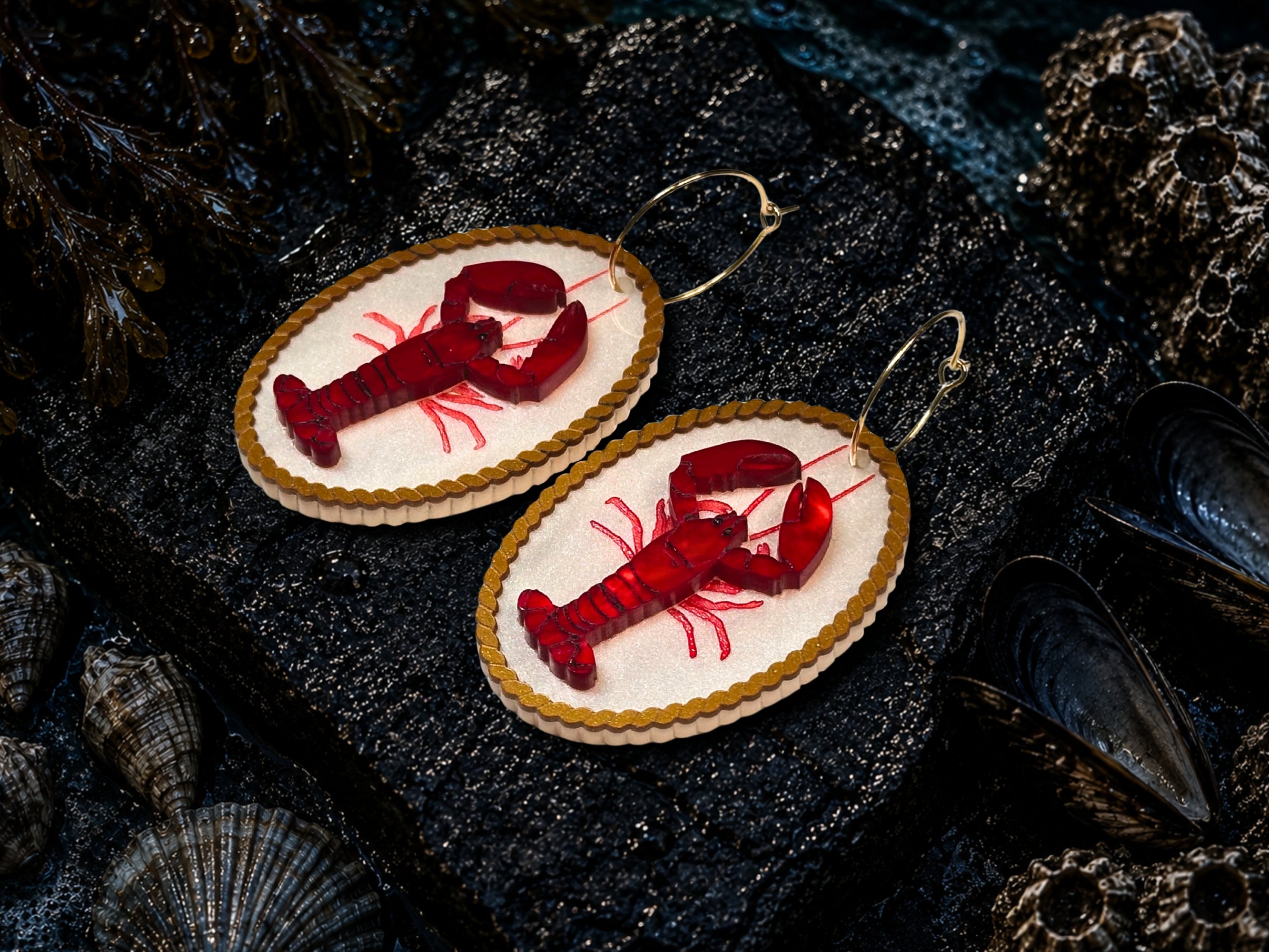Red lobster earrings photographed with shells on a dark coastal flatlay
