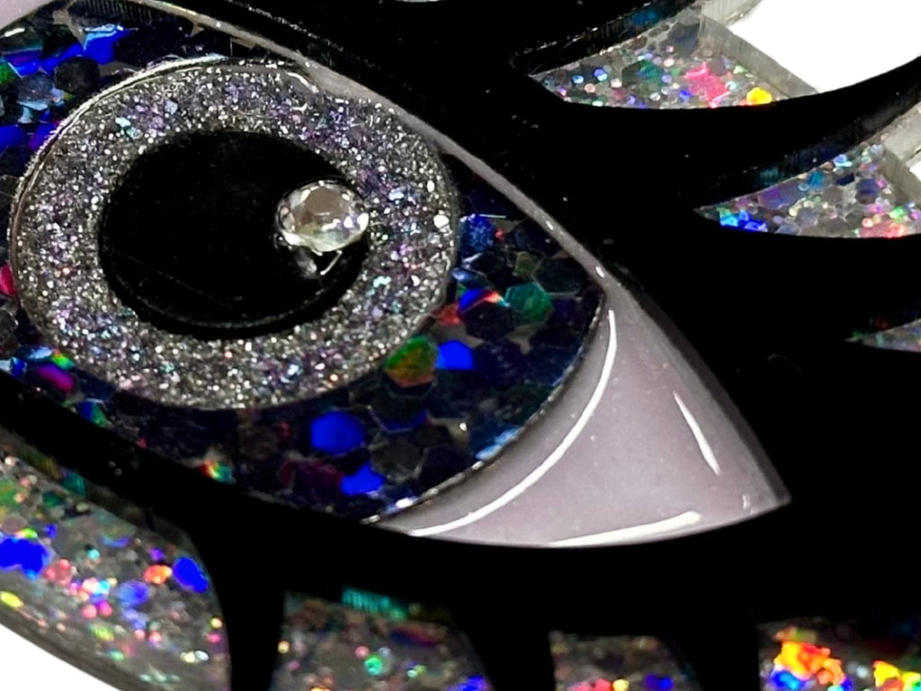 Close-up of the Siren Eye necklace showing layered glitter acrylic, resin eye details, black lashes, and crystal accent.