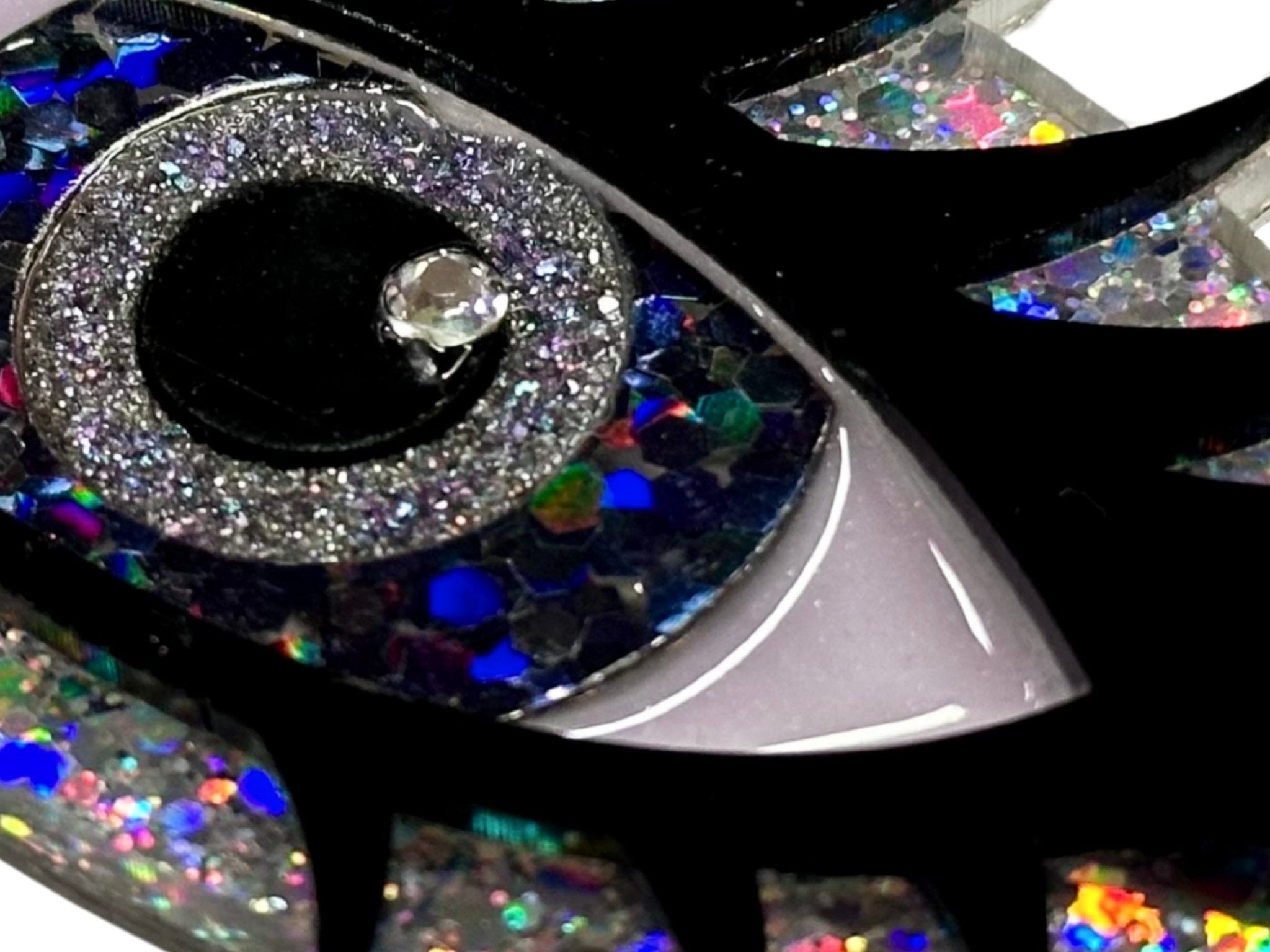 Close-up of the Siren Eye necklace showing layered glitter acrylic, resin eye details, black lashes, and crystal accent.
