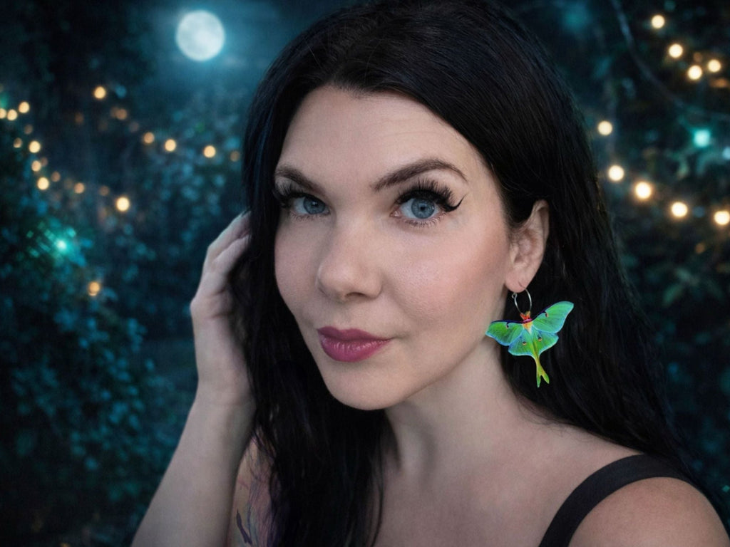 Woman with dark hair modeling oversized green luna moth statement earrings, photographed against a dreamy dark teal background with glowing lights.
