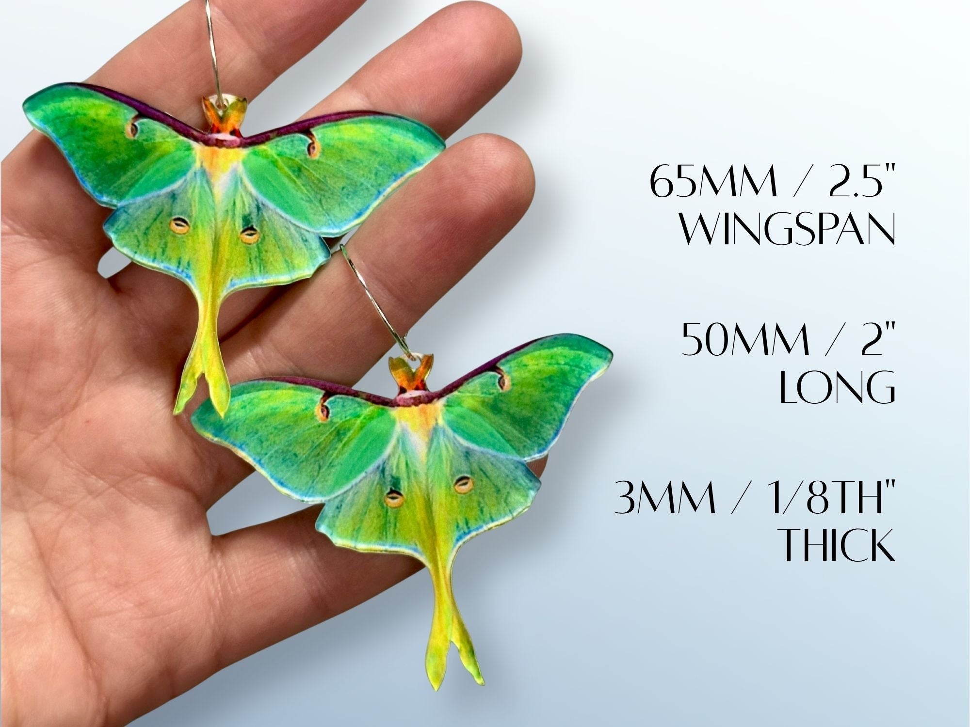 Hand holding two oversized luna moth earrings for scale, with text showing dimensions: 65mm wingspan, 50mm length, and 3mm thickness.