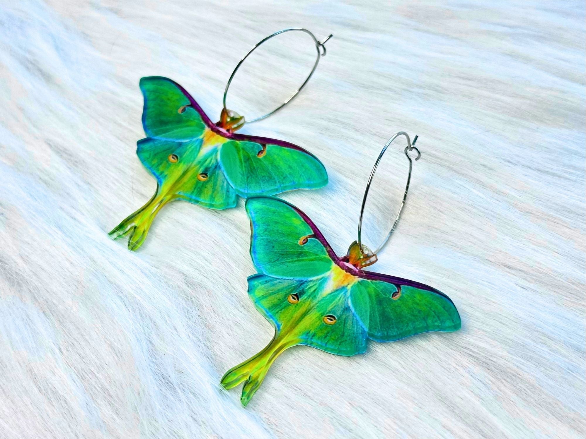 Pair of handcrafted luna moth earrings laid flat on a white fur surface, showing delicate wing details, silver hoops, and translucent green coloration.
